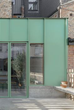 a green building with lots of windows next to a bench and some chairs on the sidewalk