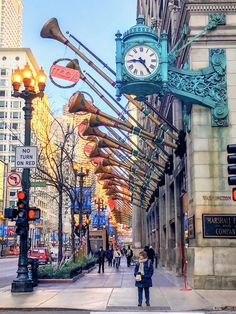 a woman standing on the side of a street next to tall buildings with clocks hanging from it's sides