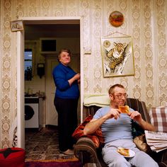 two people in a living room one is eating and the other is looking at something