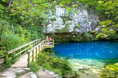 a wooden bridge over a blue pool surrounded by greenery and rock formations in the woods