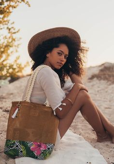 a woman sitting on the beach with her handbag in front of her, she is wearing a straw hat