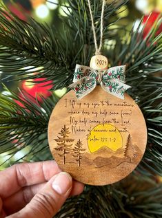 a hand holding a wooden ornament with a poem on it and pine trees in the background