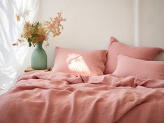 a bed with pink sheets and pillows in front of a white wall, vase with flowers