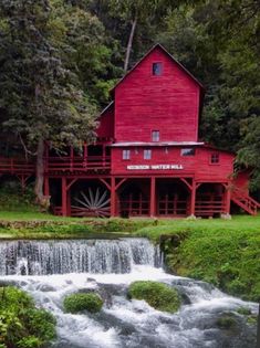 a red building with a waterfall in the foreground and green grass around it, surrounded by trees