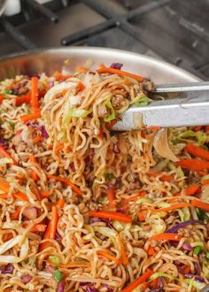 a pan filled with noodles and vegetables being stirred by tongs on the stove top