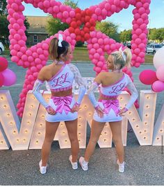 two cheerleaders are standing in front of some pink balloons and heart shaped letters