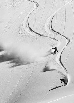 two people skiing down a snow covered slope in black and white photo with dust behind them