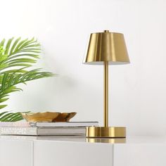 a white table with a gold lamp and books on it next to a potted plant