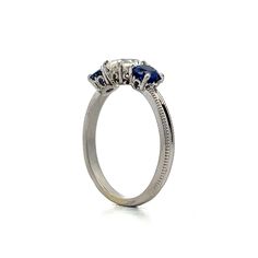 This exquisite three-stone engagement ring features a .93 carat antique cushion-cut diamond with VS2 clarity and J color. Flanked by two vibrant round blue sapphires, each weighing .46 carats, the sapphires add elegance to the overall design. The polished platinum band showcases intricate milgrain detailing. Decorative baskets cradle each gemstone, ensuring beauty from every angle. This ring blends refined craftsmanship with eye-catching details, making it a stunning choice. Luxury Three Stone Sapphire Ring, Luxury Three-stone Sapphire Promise Ring, Luxury Three Stone Sapphire Promise Ring, Three Stone Sapphire Round Ring Fine Jewelry, Sapphire Three Stone Round Cut Diamond Ring, Oval Sapphire Three-stone Diamond Ring, Oval Sapphire Three Stone Diamond Ring, Sapphire Three Stone Diamond Ring, Three Stone Sapphire Diamond Ring With Round Cut