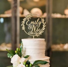 a wedding cake with white flowers and greenery on the top is decorated with an acrylic name