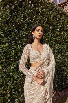 a woman standing in front of a bush wearing a white sari and matching jewelry