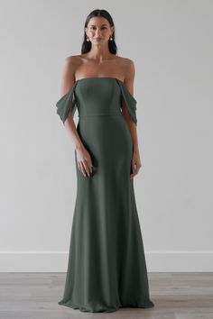 a woman wearing an off the shoulder dress with one shoulder in olive green, standing against a white wall