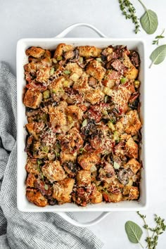 a casserole dish filled with stuffing and vegetables on a white plate next to some herbs