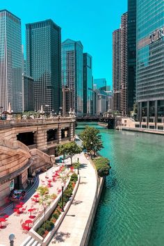 a river runs through the center of a city with tall buildings on either side and red umbrellas in the foreground