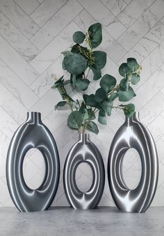 three metal vases with plants in them on a marble countertop against a white tiled wall