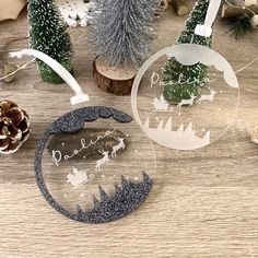 three glass christmas ornaments on a table with pine cones and fir trees in the background