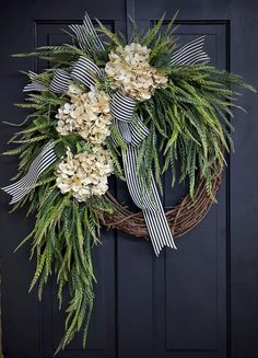 a wreath with white flowers and greenery hanging on a black front door, decorated with ribbon