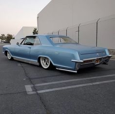 an old blue car parked in a parking lot next to a white wall and fence