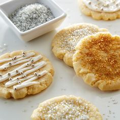 several cookies with white frosting and silver sprinkles on a plate next to a bowl of sugar