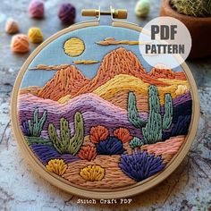 an embroidered desert scene with cacti and mountains in the background is featured on a wooden hoop