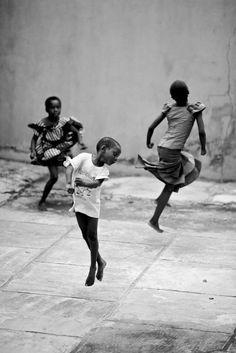 three young children are playing soccer on the sidewalk in front of a wall and building