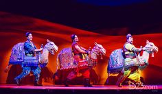 three men and two women in colorful costumes riding horses on stage with red walls behind them