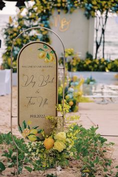 a sign with flowers and greenery on the ground next to some plants in front of it