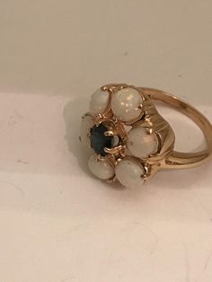 This is a beautiful cocktail ring. The ring features six opals set in a flower form fare. Material: Solid yellow gold 14k. Stamped 14k. Gems: - One synthetic sapphire. - Six preciosus white opals. The opals are slightly different. Each opals measure 5-6mm. All opals have a beautitul and soft coloring. One of them has a small spot of 1 mm and another has a blue fire inside different from the others. That asymmetry gives him personality. It is aprox. size 7 -8 (18 mm diameter). It weighs: 4.82grs. Small Vintage Ring, 1950s Rings, Vintage White Cluster Ring With Gemstone, Vintage Blue Cluster Rings, Vintage Blue Cluster Ring In 14k Gold, Vintage Cluster Sapphire Ring Gift, Luxury Vintage Blue Cluster Ring, Vintage Blue Hallmarked Cluster Ring, Vintage White Cluster Jewelry