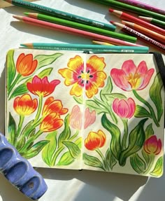 an open notebook with flowers painted on it and colored pencils next to it,