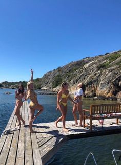 three women in bikinis are standing on a dock near the water and one woman is holding her hand up