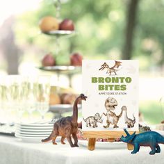 a table topped with toy dinosaurs next to a sign that says bronto bites appetizers station