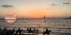 people are standing on the beach watching the sun go down over the water with a bridge in the background