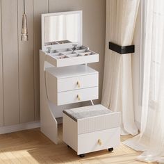 a white jewelry box sitting on top of a wooden floor next to a window with curtains