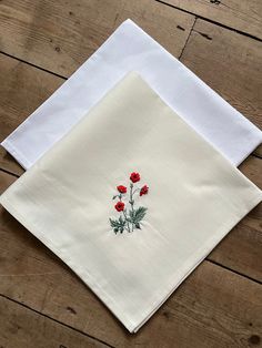 two white napkins with red flowers on them sitting on a wooden table next to each other