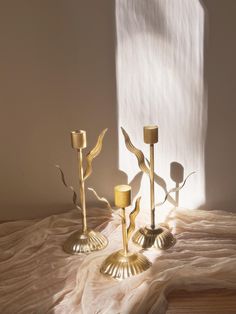 three gold candlesticks sitting on top of a white cloth covered floor next to a window