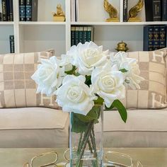 a glass vase with white roses on a table in front of some bookshelves