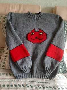 a gray sweater with a red face on it sitting on top of a couch next to a pillow