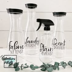 three clear glass bottles with black lids are next to some green leaves and one has a sprayer on it