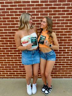 two young women standing next to each other in front of a brick wall holding books