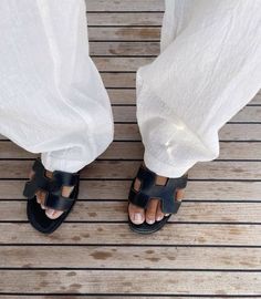 a person wearing white pants and sandals standing on a wooden floor with their feet crossed