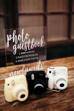 two cameras sitting on top of a wooden table next to a sign that says photo guest book