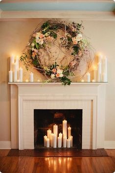 candles are lit in front of a fireplace with a wreath above it and flowers on the mantle