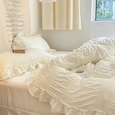 an unmade bed with white ruffles and pillows on it, next to a window