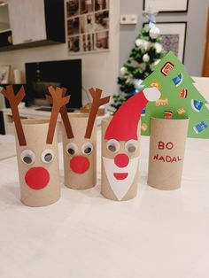 three toilet paper cups with reindeer faces and noses on the table in front of a christmas tree