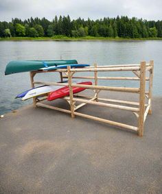 Freestanding log kayak rack on a concrete boat ramp next to a lake.  There are several kayaks and a canoe on the rack.  Several trees surround the lake in the background. Wood Kayak Rack Plans, Landscaping Blocks, Wood Kayak, Dock House, Water Vessel, Northern White Cedar, Kayak Storage, Kayak Rack, Cedar Log