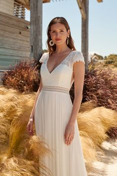 a woman wearing a white dress standing in front of some tall grass and bushes with her hands on her hips