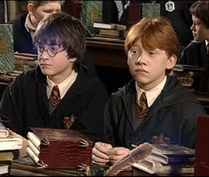 two young boys sitting at a desk with books and papers in front of them, both wearing harry potter robes