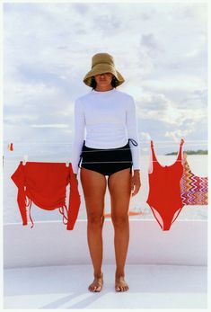 a woman standing on the beach wearing a hat and black swimsuit with her hands in her pockets