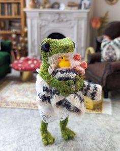 a knitted frog holding a stuffed duck in its arms, with a fireplace in the background