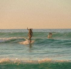 two people in the ocean on surfboards with one holding up her hand and another standing at the edge of the water